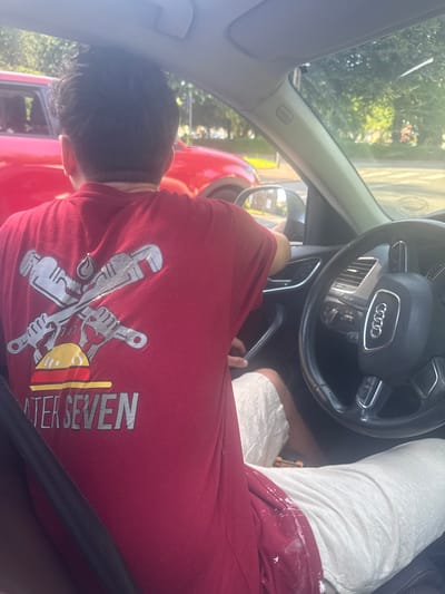 The image depicts a person in the driver's seat of an Audi. The steering wheel displays the Audi logo. The person is wearing a red t-shirt with a plumbing-related design on the back. Their legs are visible, with white shorts and a red stain, possibly paint, on one leg. The person is wearing a seatbelt. The location is Marcq-en-Barœul, France, based on the "Water Seven" t-shirt. The subject's head appears to be resting near the driver-side window.