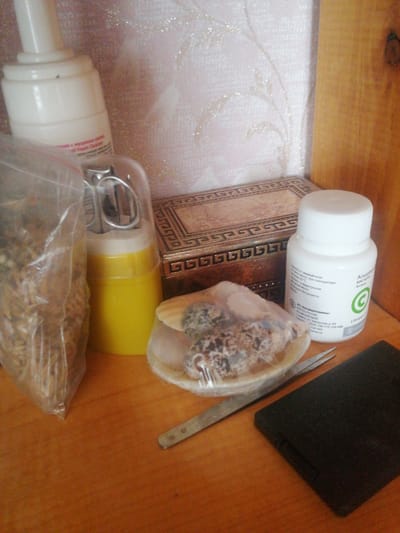 This image depicts a collection of items arranged on a wooden surface. The primary subjects are various objects, including a white pump bottle with Russian text, a small yellow container with a clear lid, a plastic bag filled with what appears to be seeds or grains, a decorative box with intricate patterns, a white bottle of what looks like vitamin supplements with Russian text and a green circular logo, and a small pair of tweezers next to a black rectangular object. The background features a textured wall with a subtle floral pattern and some sparkling elements, along with a portion of a wooden shelf on the right side.
The scene appears to be set indoors, likely on a table or shelf in a home or a similar environment. There is no discernible activity or event taking place; the items are simply arranged. The lighting suggests it is daytime, but no specific time can be determined. The visible text is predominantly in Russian, with the white pump bottle mentioning "Foam Cleanser" and "mucus" (likely referring to snail mucin in a cosmetic product), and the supplement bottle identifying the contents as "Ascorbic Acid Vitamin C" and listing the manufacturer as "Altai Vitamins." The overall impression is one of a personal collection of beauty products, supplements, and small decorative or functional items. The setting is simple and functional, with no strong emotional cues. The context of Dubrovka, Russia is provided, but there are no specific visual cues within the image to confirm or deny this location.
