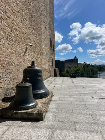 This image captures a view of a stone wall and two large bells in the foreground, with a medieval-style castle and a river in the background. The sky is a vibrant blue with fluffy white clouds, suggesting a clear and sunny day. The stone wall in the foreground is constructed from rough-hewn, tan-colored stones, laid in a rustic pattern. Two large, dark-colored bells are prominently displayed on a stone pedestal. They appear to be old and possibly decorative, with intricate designs visible around their rims.
Beyond the bells, a paved walkway made of large, light-grey stone tiles stretches into the distance. To the left of the walkway, a massive stone structure, possibly part of a fortification or historical building, rises up. Further in the background, a substantial stone castle with towers and battlements sits atop a hill, surrounded by lush green trees. A body of water, likely a river, is visible to the right of the castle, with some structures on the opposite bank.
The overall scene evokes a sense of history and grandeur. The bright daylight and blue sky contribute to a pleasant and inviting atmosphere. The presence of the castle and the ancient bells suggests a historical site or museum. While no people are visible in the image, the well-maintained walkway and the presence of what appears to be a security camera on the stone wall indicate that this is an accessible public space. The orientation of the image is such that it appears to be taken from a low angle, looking upwards towards the castle and sky.