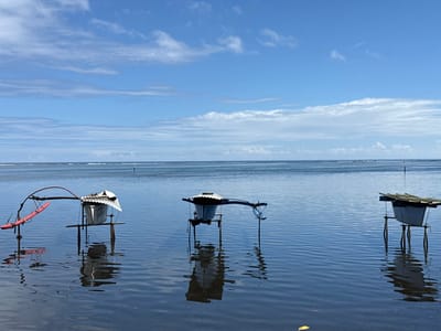 The scene is located in Māhina, France, depicting three man-made structures submerged in calm, shallow blue water under a partially cloudy blue sky. The structures, likely fish traps or aquatic farming platforms, are arranged horizontally across the midground. The leftmost unit features a white, bucket-like container topped with a corrugated metal roof, supported by dark vertical poles, and equipped with a red outrigger-style arm extending to the left via curved metal rods. The central unit is similar, presenting a white container with a corrugated metal roof and dark supports. The rightmost unit consists of a light-colored rectangular box with a flat dark roof, also supported by dark vertical poles. All structures cast clear reflections on the water's surface. The water extends to a distant horizon with subtle wave patterns. No human figures are visible.