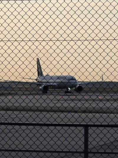 This is a photograph of an airplane on a tarmac, viewed through a chain-link fence. The airplane is a narrow-body jet, predominantly white with some gray detailing. It is positioned on a runway, facing left, with its engines visible. The tail fin is prominent and appears to be dark gray. Red and blue lights are visible on the tarmac. The sky is a hazy, pale orange, suggesting either sunrise or sunset. The foreground is dominated by the black chain-link fence, which creates a diamond pattern across the image, obscuring parts of the scene. The location is identified as near Frankfurt Airport (FRA) in Kelsterbach, Germany, a major international airport serving the Frankfurt Rhine-Main region. The image captures a moment of stillness before takeoff or after landing.