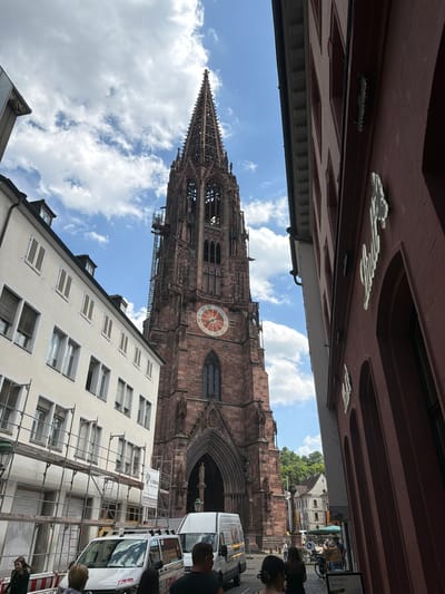 The image shows a street view in Freiburg im Breisgau, Germany, with a prominent Gothic cathedral dominating the scene. The cathedral, constructed of red sandstone, features a tall, ornate spire and a large circular clock face with Roman numerals. The sky is blue with scattered white clouds, suggesting a pleasant day, likely midday given the bright sunlight.
In the foreground, a light-colored building with multiple windows is partially obscured by scaffolding, indicating ongoing construction or renovation work. Several white vans are parked on the street, one of which has visible text that appears to be promotional or informational, though it is mostly unreadable.
Several people are visible, walking or standing on the sidewalk. Their presence suggests the area is a public space, possibly a popular tourist destination. The perspective of the photograph is from a low angle, looking upwards towards the cathedral and the buildings, creating a sense of grandeur and scale. The architecture of the buildings and the style of the cathedral are characteristic of European city centers. The overall mood is one of urban exploration and appreciation of historical architecture.