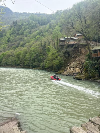 The image shows a red inflatable boat in a river at Kveda Makhuntseti, Georgia. The boat is traveling rapidly through the water, creating a white wake behind it. On the left bank of the river, there's a dense forest covering a steep hillside. Higher up the hill are a few wooden buildings constructed on stilts, nestled amongst the trees. The river's water is a murky green color. The right bank is rocky. The sky is overcast. Overhead power lines are visible.