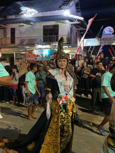 This nighttime image captures an outdoor cultural event in Desa Kalida, RT 12 RW 04, Kedungbanteng, Indonesia.

The central subject is an individual, appearing female, adorned in elaborate traditional attire, positioned slightly right of the frame's center. She wears a golden, ornate headpiece with a dark, possibly feathered, upright element. Her main garment is a black and gold ensemble featuring intricate patterns, with a long, dark train extending behind her. A prominent sash crafted from folded Indonesian Rupiah banknotes is draped over her left shoulder and across her chest, secured near her waist with colorful ribbons and decorative elements. She has makeup on her face and is smiling, looking directly towards the viewer.

Behind and around the central figure, approximately twenty-five to thirty spectators are visible, observing the scene. They are dressed in casual clothing, including t-shirts, hoodies, and hijabs, standing and conversing. Some individuals are partially obscured by the central figure or other members of the crowd.

The background features a two-story building with a light-colored, tiled roof, visible windows, and a balcony railing. To the right, a metal archway structure is present, displaying an eagle logo with the text "LPDH BANK" and "DESA KALIDA RT 12 RW 04". An Indonesian flag (red and white) hangs vertically from this structure. String lights are visible in the upper right background, contributing to the night illumination. The ground surface is paved, and artificial lighting casts distinct shadows, indicating a well-lit outdoor setting after dark.