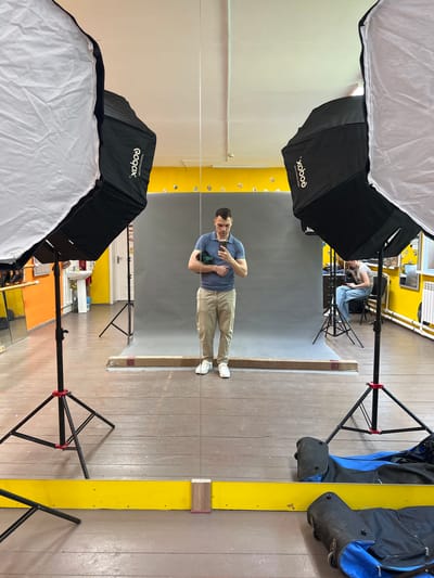 The image depicts a mirrored view of a man standing in what appears to be a photography studio in Bolshiye Kabany, Russia. The man is wearing a light blue short-sleeved polo shirt, tan pants, and white sneakers. He is holding an object in his hands, possibly a camera or phone, pointed towards himself. The background is a grey screen stretched on a frame. Above and below, there are two large black and white softboxes on stands, positioned to provide lighting for a photoshoot. Behind the grey screen, there are signs of a cluttered workspace with yellow-painted walls and various equipment. Another person is sitting in the background, near a computer. The floor is made of wooden planks. There is a narrow yellow board attached to the floor on the left and right. The presence of lighting equipment indicates the location is likely a professional or amateur photography studio.