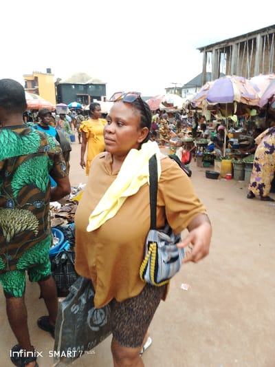 A bustling outdoor market scene showcases an assortment of people and goods. A woman with sunglasses perched on her head stands in the foreground, wearing a brown shirt and patterned shorts, with a handbag slung over her shoulder and a yellow cloth draped on her shoulder. Behind her, a man in a vibrant green patterned shirt and shorts is visible. Further in the background, numerous vendors are stationed under colorful umbrellas, displaying their wares to potential customers. A building is visible in the background.