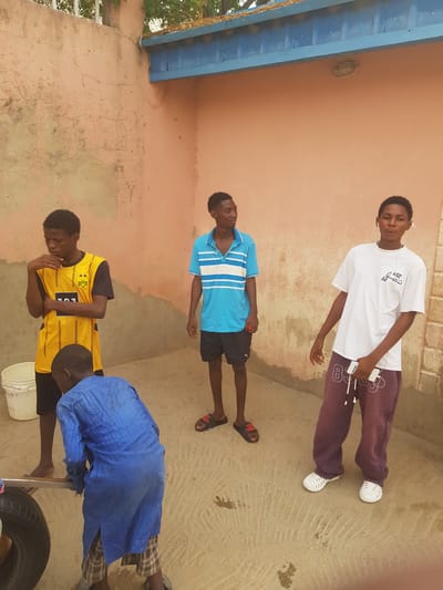 The image shows a group of four young men standing in an outdoor space in Dutsinma A, Nigeria. One is wearing a white t-shirt, maroon sweatpants and white shoes. The second individual is wearing a light blue polo shirt with white stripes and black shorts. The third is in a yellow soccer jersey. The fourth man, wearing a dark blue coverall, appears to be working on a car tire with a metal tool. A white bucket is near the tire. They are positioned on a concrete surface adjacent to a peach-colored wall. A blue awning is partially visible in the background.