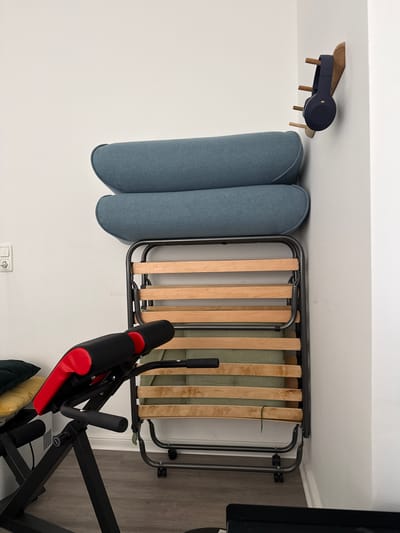 The image shows a room with a white wall and a wooden floor. In the center of the image is a foldable metal bed frame with wooden slats. To the left of the bed frame are two upholstered cushions, likely part of a sofa or seating area, in a muted blue-grey color. Above the cushions, mounted on the wall, is a small, dark blue, modern wall shelf with wooden dowels, possibly designed to hold headphones or other items. To the right of the bed frame is a black and red exercise machine, possibly an ab roller or a similar piece of home fitness equipment. The scene appears to be indoors, likely in a residential setting in Wilhelmshaven, Germany. There is no indication of the time of day or weather. The lighting is neutral, suggesting daytime indoor lighting. The overall impression is a slightly cluttered but functional living space. There is a white electrical outlet visible on the wall near the floor.
