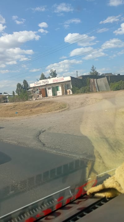 A street-level view shows a "Glory Paints" shop with the tagline "Make a Lasting Impression" under a partly cloudy sky. The shop sits on a corner, bordered by dusty dirt roads and some vegetation. Corrugated metal sheets stand adjacent to the shop. Part of the vehicle's dashboard is visible in the foreground, with some red accents and a light-colored cloth. Overhead wires stretch across the sky.
