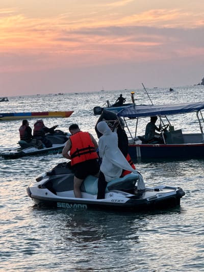 The image depicts an active water recreation scene at Cenang Beach, Malaysia, under a sky displaying pink, orange, and light blue hues characteristic of either sunrise or sunset.

In the foreground, a white and black "Seadoo GTX" jet ski is positioned on moderately choppy water. A male individual, wearing an orange life vest over dark shorts, is seated on the forward section of this jet ski. Behind him, a female individual, wearing a light-colored head covering (hijab) and a long-sleeved light-colored garment over dark pants, is seated, with her right arm raised and extended. To the left and slightly behind, a second "Seadoo GTX" jet ski holds two additional individuals, both wearing dark clothing and life vests, facing towards the left.

To the right of the foreground jet ski, a blue and white motorboat equipped with an overhead canopy is visible. A male individual, wearing a patterned shirt, is seated at the helm, operating the vessel. In the middle-ground to the far left, a yellow and red inflatable watercraft is partially visible. Further into the background, a dark silhouette of a person stands in a smaller boat featuring an outboard motor and a pole with an orange light. Numerous other small boats are scattered across the water towards the distant horizon, where a sailboat is also discernible.