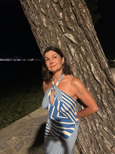 A woman with brown hair and fair skin stands next to a large tree trunk at night. She is wearing a blue and white striped halter dress. Her hands are behind her back. In the background, there is water, a stone pathway, and a distant city skyline illuminated by lights. The location is the city of Port De Pollença, Spain.