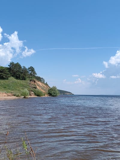 A wide shot captures a serene landscape featuring a calm body of water under a clear blue sky dotted with white clouds. The foreground is dominated by the dark, rippling water, with the gentle waves reflecting the sky. Along the left side of the image, a sandy shore leads to a grassy embankment covered with green vegetation and scattered trees, some of which are pine trees. The embankment rises to a cliff face with exposed soil, showing layers of earth. In the distance, a long, low-lying hill stretches across the horizon. The scene appears to be a natural, undeveloped area, possibly a riverbank or a lake shore, in the vicinity of Panskaya Sloboda, Russia. There are no people or animals visible in the image. The overall impression is one of peaceful tranquility and natural beauty.