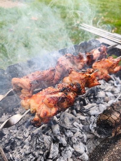 The image shows two skewers of meat, likely chicken, grilling over hot coals in an outdoor setting, likely in Орлиновский округ, Ukraine. Smoke is rising from the coals and the meat, indicating it is being cooked. The coals are a mixture of gray ash and some larger pieces of burned wood. In the background, there is green grass. A grill rack is visible at the top.
