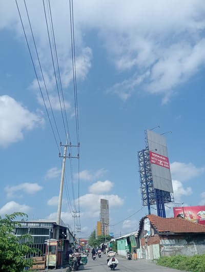 The image depicts a street scene in Sawotratap, Indonesia, under a bright blue sky with scattered clouds. Overhead, power lines stretch across the frame, supported by a concrete utility pole on the left. On the right side of the street, a tall, multi-story billboard structure stands, partially covered by a grey tarp. A red banner on the billboard displays "DISEWAKAN:" followed by two phone numbers. Adjacent to the billboard is a weathered building with a red-tiled roof. Further down the street, several buildings with corrugated metal roofs line the road. A few individuals are present, some riding motorcycles, suggesting ongoing activity. A food stall, possibly selling "Sate Padang," is visible on the left side of the road. The overall impression is a busy street with a mix of commercial and residential elements in an Indonesian urban or semi-urban setting.