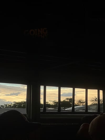 The image depicts a view from an interior space looking out through a window or opening at a sunset. The foreground is dominated by dark, silhouetted shapes, likely furniture or people, suggesting the viewer is inside a building or vehicle. Above these shadows, the window reveals a sky with a gradient of warm colors, transitioning from a pale blue and white with scattered clouds to a strong yellow-orange hue near the horizon. Silhouetted trees and structures are visible against this sky. The window is framed by dark, vertical bars, creating a grid-like effect over the outdoor scene. In the upper part of the image, within the interior darkness, there are yellow illuminated letters that read "GOING". The scene appears to be set in El Guasaule, Nicaragua, as indicated by the prompt's context.