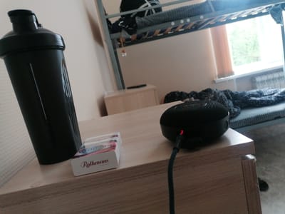 A black plastic water bottle shaker and a pack of Rothmans cigarettes sit on a wooden nightstand. A small, black, round speaker is also on the nightstand, plugged into a charging cable. The nightstand is positioned in a bedroom with a bunk bed and a window visible in the background. The overall scene suggests a personal living space. The location is Komsoomolskiy, Russia.