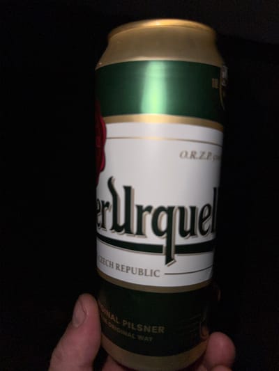 A close-up, eye-level shot shows a person's thumb and forefinger holding a can of Pilsner Urquell beer. The can has a white label with "Pilsner Urquell" in large, dark green lettering. The top of the can is gold and green. The background is dark and out of focus. The scene appears to be indoors.