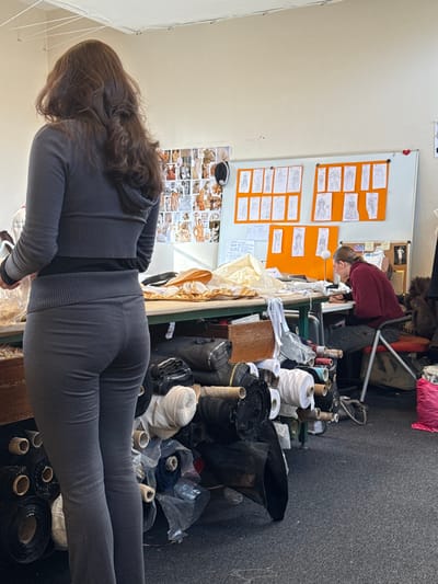 This image depicts an indoor workshop or design studio located in Arcueil, France. The scene features two individuals and numerous items related to garment design or production.
In the foreground, slightly to the left of the center, a female subject stands with her back predominantly to the viewer, facing towards the right side of the frame. She has long, dark brown, wavy hair. She wears a dark grey long-sleeved top with a ribbed waistband and matching dark grey fitted trousers. Her right arm is slightly lifted, positioned over a work surface.
To the right, in the background, a second individual, appearing male, is seated at a table, facing towards the left. He has short dark hair pulled back. He wears a dark red long-sleeved top and dark trousers. He is leaning forward, focused on items on the table in front of him. He is seated on a chair with a red back and an orange seat, upon which the word "ARCUEIL" is partially visible.
The room has light-colored, possibly off-white, walls and a dark grey carpeted floor. Across the ceiling, white electrical wires are visibly strung. Along the back wall, to the left of the seated individual, a large white whiteboard displays several sheets of orange paper, each containing multiple hand-drawn fashion sketches of figures in various poses and attire. To the left of the whiteboard, a collage of smaller images, likely reference photographs or mood board elements, is affixed to the wall.
A large, light-colored work table stretches across the mid-ground. Its surface is covered with folded cream or gold-colored fabric and various white strips of material. Beneath this table and on the floor in the foreground, numerous rolls of fabric are stacked and piled. These rolls vary in color, including black, white, cream, blue, and dark grey, with some wrapped in clear plastic. A wooden box is also visible among the fabric rolls under the table. To the far right, on the floor, a pink plastic bag is present. The space is illuminated by overhead lighting and ambient light from an unseen source.