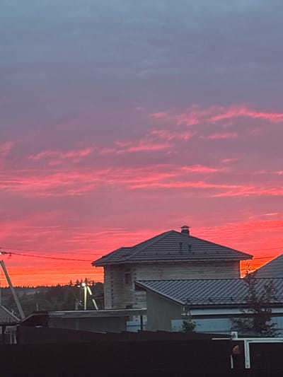 The image depicts a sunset scene in the city of Dolce Vita KP, Russia. The sky is a vibrant gradient of pink and orange, with thin, elongated clouds streaked across. In the foreground, the rooftops of several houses are visible, constructed with dark grey tiles. One prominent house, partially visible on the right, appears to be made of light-colored wood. A dark fence runs along the bottom of the frame, obscuring the lower portions of the structures and the ground. In the distance, a line of trees can be seen against the horizon, with utility poles and wires interspersed. The overall impression is of a residential area at dusk.