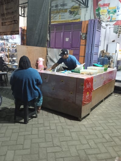 At ground level, a vendor with a blue shirt and a black baseball cap is seen from the side and behind, leaning over a food stall. The stall has a pink and brown counter wrapped in a patterned fabric. Two clear plastic containers are on the counter, one holding some food. To the left, a person wearing a blue sweater and a black head covering is seated on a dark stool, facing away from the camera. The background features a purple door, a large banner with text, and shelves filled with items. The ground is paved with grey rectangular blocks. The scene appears to be outdoors, likely at night due to the lighting. The image is taken in Cangkring, Indonesia.