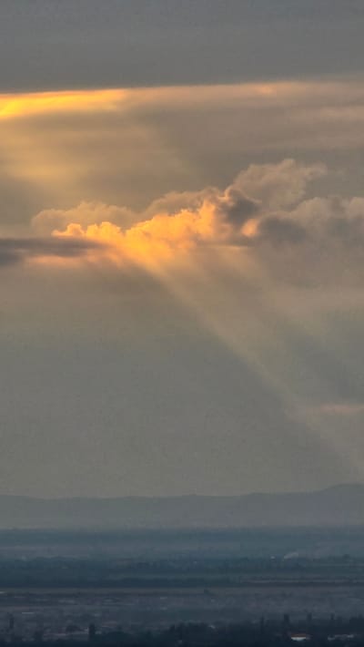 The image shows a dramatic sky with sunbeams breaking through clouds, casting a warm, golden light. The sunbeams are prominent and create a sense of depth and atmosphere. Below the sky, there is a hazy, layered landscape that appears to be a distant city or town. The foreground is darker and suggests the presence of buildings and possibly vegetation. The overall impression is one of natural beauty and a serene, possibly twilight, setting. The location is identified as Alkhazurovo, Russia, implying a view over this area during a specific time of day when the sky displays these characteristics.