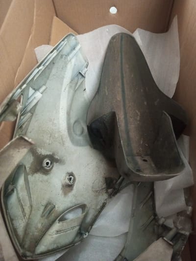 A cardboard box contains disassembled scooter parts. The components, primarily gray and dirty, include a headlight cover with two screw holes, a dark fender, and other fragmented pieces. White protective tissue is interspersed among the parts. The overall appearance suggests used or damaged scooter components.