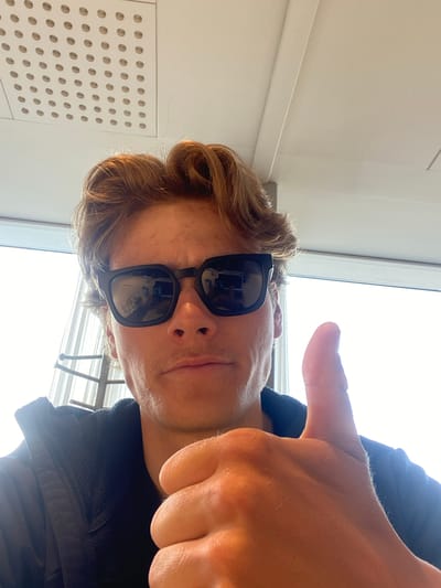A person with light skin and brown hair is wearing sunglasses and giving a thumbs-up. They are indoors, next to a window. To the left is a white wall with circular perforations. The location is Kastrup, Denmark.
