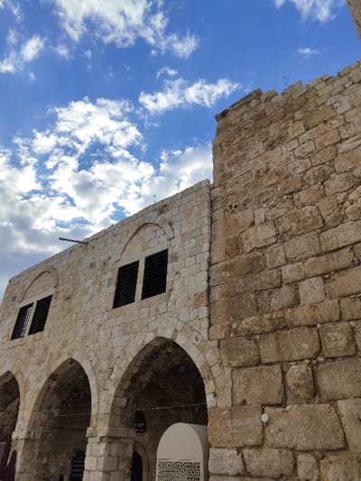The image displays a historic stone building in Rosh HaAyin, Israel, under a blue sky with scattered white clouds. The structure is composed of light-colored, irregular stone blocks, showing signs of age. Its lower section features a series of pointed arches, forming an arcade. The upper facade includes rectangular windows fitted with black grates. The right side of the building exhibits larger, more weathered stone blocks and a jagged, partially ruined top edge from which small vegetation grows. A dark conduit or pipe protrudes from the upper-left area of the structure. Below one of the arches, a small white structure with a decorative base is present, alongside a sign displaying the Hebrew text 'מבוא ב' (Mavo Bet), translated as 'Entrance B'. No individuals are depicted within the frame.