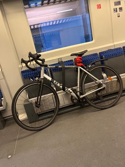 A light grey and black road or gravel bicycle, featuring "BTWIN" on its frame, is mounted horizontally inside a train carriage. A red bag is attached to the bike's saddle, and the bicycle is secured by a black strap around its frame. Below the bike, blue patterned train seats are visible, some appearing folded, along with an accessibility symbol (wheelchair icon) on the bike rack structure. To the left, a large window reflects a dark, blue-tinged exterior, suggesting the scene takes place at dusk or night. The train's interior walls are light-colored, displaying a red symbol with the number "2" and a separate red square with the number "2", likely denoting a second-class carriage. The floor is grey with a speckled pattern. The image captures the practical transport of a bicycle within a train, consistent with travel in Germany.
