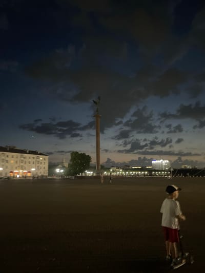 This is a medium-shot, eye-level, outdoor photograph taken at dusk or early night. The scene depicts an open, paved area with a prominent monument on the right side, a large building on the left, and a cityscape in the background.

In the foreground, a young boy, appearing to be Caucasian, is standing to the right of the frame, facing towards the left. He is wearing a white t-shirt, red shorts, a baseball cap, and what appear to be sneakers. He is standing next to a scooter, with his hands resting on the handlebars. His body is angled slightly away from the viewer.

The middle ground features a vast, dark paved surface, possibly a plaza or square. Further back, on the left side of the image, is a large, light-colored building with illuminated windows, suggesting it is occupied or in use. Adjacent to this building, a tall, slender monument with a statue at its apex rises into the sky. Behind the monument, trees are visible. Across the expanse, a line of streetlights illuminates the distance, with more buildings forming the cityscape. The sky is dark blue, with scattered clouds that are tinged with lighter colors from the fading daylight or artificial lights. The overall lighting is dim, creating a twilight atmosphere.

The location is identified as Verkhnyaya Stavropol'ka, Russia.
