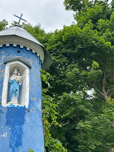 A weathered, blue-painted cylindrical structure stands against a backdrop of lush green trees under a cloudy sky. The structure has a conical metal roof topped with a blue cross. A niche in the structure features a statue of the Virgin Mary holding the infant Jesus. The statue is adorned with a blue robe and a golden crown. The blue paint on the exterior of the structure is peeling, revealing white underneath. Green vines climb the side of the structure. The location is in Хаценчыцкі сельскі Савет, Belarus.
