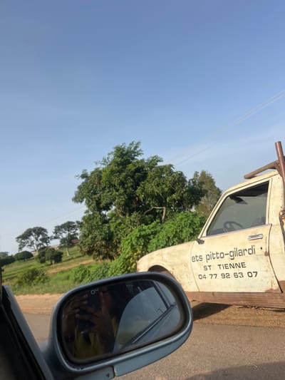 The image shows a side view of an old, weathered white truck with text on its door that reads "ets pitto-gilardi", "ST TIENNE", and "04 77 92 63 07". The truck appears to be parked on a dirt road with a grassy embankment and trees in the background. A side mirror of another vehicle is in the foreground, reflecting the sky and a person holding a phone. The sky is clear and blue. The environment suggests a rural or semi-rural setting, consistent with the outskirts of Ladura Foron, Nigeria.