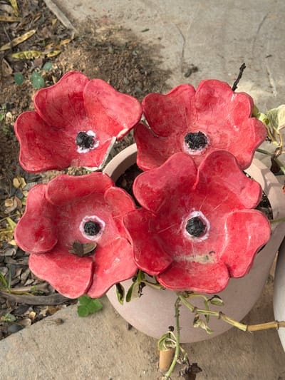 A top-down view features four ceramic red poppies with black centers nestled in a clay pot. The textured, weathered petals reveal subtle variations in color and cracks, adding a rustic charm. The pot is surrounded by dried leaves, green foliage, and a concrete surface, suggesting an outdoor setting.