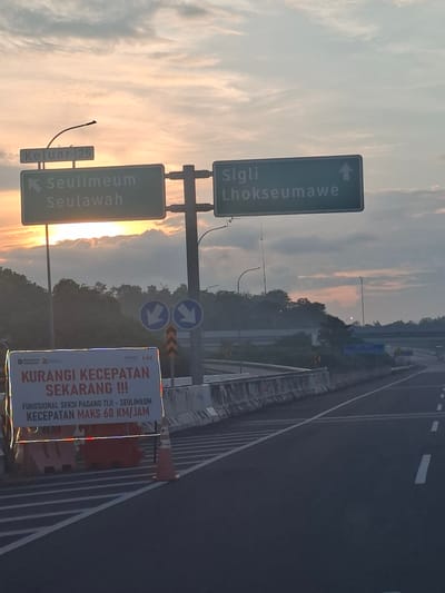 An outdoor, eye-level, medium shot captures a highway scene in Lambarotunong, Indonesia, at dusk. The sky is filled with a soft palette of peach and gray clouds, illuminated by the setting sun. Prominently displayed are highway signs: one indicates "Keluar 25" (Exit 25) with an arrow pointing left, and another green sign shows "Seulimeum Seulawah" with a left-pointing arrow. To the right, a similar green sign reads "Sigli Lhokseumawe" with an upward-pointing arrow. Below these, two blue circular signs with white arrows pointing downwards are visible.
In the foreground, a red and white banner is erected on an orange barrier, displaying text in Indonesian. The banner reads "KURANGI KECEPATAN SEKARANG !!!" (REDUCE SPEED NOW !!!) and further specifies "FUNGSIJSIONAL SEKSI PADANG TJIJI - SEULIMEUM KECEPATAN MAKS 60 KM/JAM" (FUNCTIONAL SECTION PADANG TJIJI - SEULIMEUM MAX SPEED 60 KM/HOUR). Traffic cones and more orange barriers line the side of the highway, which has clear lane markings. The highway stretches into the distance, curving to the right, with overpasses visible further along. The surrounding environment is characterized by dense greenery on a hillside. The lighting suggests it is twilight, creating a somewhat hazy atmosphere.