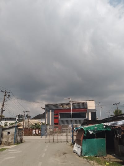 The image displays a street scene in Mgbuoshimini, Nigeria, under a heavily overcast, dark grey sky. A paved road with a faded white centerline extends from the foreground towards the mid-ground. To the immediate right of the road, a makeshift stall is constructed from reddish-brown corrugated metal sheets and a green tarp. On the left side, a small, light-colored concrete building with a rectangular window is visible, followed by several wooden utility poles equipped with multiple power lines and cross arms.

In the mid-ground, a modern building is prominent, identified by gold lettering on its facade as "THE THIRD AVENUE EVENT PLACE." This structure features a central red section with large reflective windows, bordered by vertical grey and white architectural elements. Two white streetlights on poles are positioned in front of the building. A large, partially open metal gate, comprising multiple grey vertical bars, restricts access to the event place. Behind this gate, a single individual dressed in light-colored clothing is visible, walking away from the camera. Posters are affixed to a fence situated behind the gate. The background reveals additional residential structures and varying densities of green vegetation.