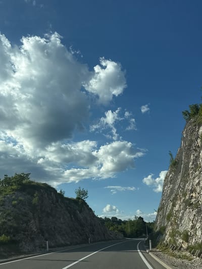 This image depicts a road cutting through a rocky, mountainous landscape. On the left, a steep, dark, rocky incline is covered with sparse vegetation. On the right, an equally steep, light-colored, rocky cliff face rises dramatically, with small plants clinging to its surface. A paved road with white lane markings curves through the scene, bordered by low concrete barriers and reflective markers. The sky is a vibrant blue, filled with dramatic, fluffy white clouds, some of which cast shadows on the landscape below. Lush green trees and foliage are visible in the distance, suggesting a natural, undeveloped environment. A solitary tree stands on the crest of the left-hand incline. The scene appears to be located on the road between Cetinje and Budva, near Očinići, Montenegro, a region known for its rugged terrain and scenic beauty. There are no people visible in the image.