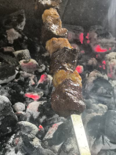 A close-up, vertical shot shows a metal skewer with several pieces of grilled meat over glowing coals. The meat appears to be well-cooked, with some pieces showing a darker, charred exterior and others a lighter, browned surface. The coals are a mix of black and white ash, with several sections emitting a bright red glow, indicating intense heat. The background is out of focus and appears to be composed of more coals and smoke. The scene suggests an outdoor cooking environment, likely a barbecue or grill. The image does not contain enough information to determine the specific location in Bina, Azerbaijan.
