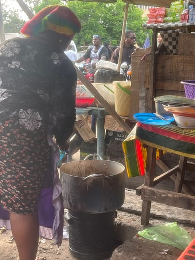 The image captures an outdoor market or street stall scene during daylight hours. The perspective is from behind a person in the immediate foreground, looking towards the stall and other individuals.
In the foreground, a person is seen from the back, wearing a dark-colored garment with a white floral or abstract pattern. They have a distinctive, colorful hat or head covering with horizontal stripes in hues of red, orange, yellow, green, and blue/purple. A purple fabric is visible to their right (viewer's left), possibly part of their attire or an item they are carrying. The ground appears to be a mix of dirt and concrete.
To the right of the foreground person, a prominent black, cylindrical machine or large pot stands on the ground. It has a metallic (silver/grey) funnel-like top or lid and a handle. This object suggests some form of food preparation or processing is taking place at the stall.
The midground features a rustic wooden stall structure with shelves and support beams. Various items are displayed or stored on and around it. On the upper parts of the stall, there are purple plastic baskets, white or cream-colored bowls, and a blue plastic lid. A piece of striped fabric, possibly depicting Pan-African colors (red, yellow, green, and black) or a Kente-style pattern, hangs from the stall. Below, other containers, including what appears to be a light yellow-green plastic bucket and a red container, are visible.
Several people are in the midground and background, interacting with the stall or passing by. One man, wearing a dark shirt and sunglasses, is seated on what appears to be a motorbike. Behind him, another man in a white sleeveless top with a pattern, possibly a jersey, is also visible. Both are looking towards the right side of the frame. Green foliage from trees or bushes is visible in the background, reinforcing the outdoor setting.
Visible text is limited and partially obscured. On a cardboard box located underneath the stall, the word "BUTTERS" is partially legible, with "OIL" or "SOIL" possibly below it. Another box shows partial letters "D" and "S".
The scene conveys the bustling atmosphere of a local market or roadside vendor, characterized by makeshift structures, various goods, and everyday activity.
