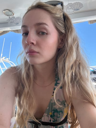 A young woman with light blonde hair and fair skin is taking a selfie on a boat. She is wearing a floral print dress. Sunglasses are perched on her head. The background shows a clear blue sky and parts of the boat's rigging and structure. A seashell design is visible on a white surface, indicating the boat's interior. The location appears to be coastal, potentially in Unknown, Mexico, inferred from the nautical setting and clear sky.