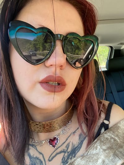 A woman with auburn hair and heart-shaped sunglasses is lying down, wearing a gold collar necklace and a silver necklace with a red pendant. She has tattoos on her neck and chest. The reflection in her sunglasses shows a street with trees and a vehicle, potentially indicating a location in Kruče - Kërruçi, Montenegro. A vehicle interior is visible in the background.