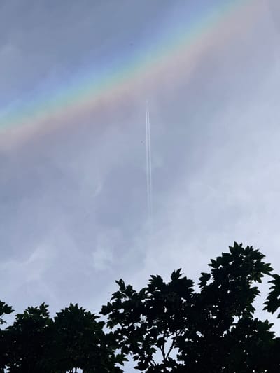 The image depicts a cloudy sky over a silhouette of tree foliage in Amstelveen, Netherlands. A faint rainbow arcs across the upper left portion of the sky. A jet plane is visible in the upper center, leaving a distinct contrail. The trees, silhouetted against the brighter sky, are dark green and detailed with visible leaves and branches. The overall lighting suggests it is daytime, possibly after or during a rain shower, given the presence of the rainbow and clouds.