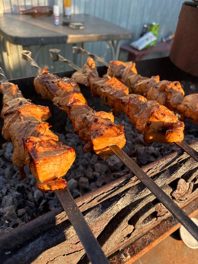 The image shows several skewers of meat cooking over hot coals in a grill. The meat is browned and appears to be chicken or pork, cut into cubes. The grill is made of metal and is filled with hot, gray coals. In the background, there is a table with bottles and containers on it. The scene is outdoors, with sunlight casting shadows on the grill and the meat. The location is the village of Dunilovo, Russia.