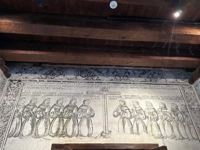 This image depicts a detailed mural on the interior wall of the former Augustinian convent of San Miguel Arcángel in Huejotzingo, Mexico. The mural is executed in a monochromatic style, primarily in black ink or paint, against a light-colored, textured wall.

The central section of the mural displays a group of Franciscan friars, distinguished by their hooded robes and ropes. They are depicted in various stages of prayer, with their hands clasped in front of them and their heads bowed. The friars are arranged in two distinct groups, separated by a large, wooden cross. The group on the left appears to be larger, with at least seven figures visible, while the group on the right has five figures. Above and between the friars and the cross are lines of text in Spanish, likely inscriptions identifying the individuals or providing context for the scene. Decorative elements, including stylized floral motifs and geometric patterns, frame the upper portions of the mural. The overall scene evokes a sense of religious devotion and historical narrative. The wooden beams of the ceiling are visible above the mural, indicating the age and architectural style of the building.
