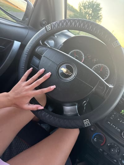 A first-person perspective shot shows a woman's left hand resting on the steering wheel of a Chevrolet vehicle. The steering wheel is black with a quilted texture and adorned with rhinestones on the left side. The woman has light-skinned hands with neatly manicured, light pink nails. The interior of the car is visible, including the dashboard with the Chevrolet logo on the steering wheel, instrument cluster, and part of the center console with climate controls. Outside the car, through the driver's side window and side mirror, a blurry background suggests an outdoor environment with trees and a sunset or sunrise. The overall impression is a snapshot taken inside a car, possibly while stopped or driving, focusing on the driver's hand and the car's interior. The location is Iordanovca, Moldova.