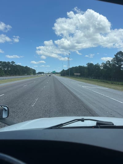 This is a POV shot from the interior of a vehicle, looking out at a highway in Shellman Bluff, Georgia, United States. The road is asphalt with white lane markers and appears to be a multi-lane highway with cars driving in the distance. To the left of the road is a grassy median and then a line of trees. To the right of the road, there is also a grassy area with trees and a sign that reads "Evergreen Villa Anderson St Evergreen Interchange". The sky is blue with scattered white clouds, and there are several tall cell towers visible in the distance. The front windshield and dashboard of the vehicle are visible in the foreground.