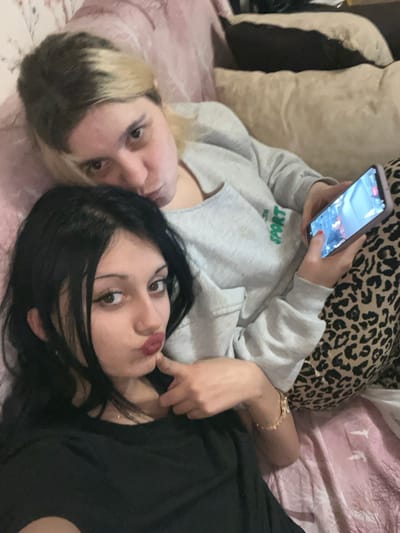 The image displays two young adult females in an indoor residential setting, consistent with a bedroom or living room environment within Lanchkhuti, Georgia. The precise location within the city is not discernible from the visual information.
In the foreground, one female with dark hair, estimated to be in her late teens or early twenties, is positioned closer to the camera. She wears a black t-shirt and looks directly at the viewer with pursed lips, her right hand gently touching her chin. A delicate gold-colored bracelet is visible on her left wrist.
Behind her, a second female, also appearing to be a young adult, with blonde hair showing dark roots, is situated. She wears a light gray long-sleeved sweatshirt, featuring a partial green "SPORT" emblem on the chest, and leopard print pants. Her left hand holds a light-colored smartphone, positioned vertically, with its screen actively displaying what appears to be a video game or application interface. She gazes towards the camera with a neutral to slightly pursed expression.
Both individuals are arranged as if posing for a selfie. They are resting on a surface covered by a light-colored, possibly pink, blanket or bedding. Additional pillows or cushions are visible in the background, one appearing light pink/white and another darker brown/beige.