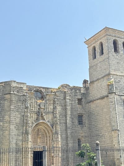 An angled perspective shows the exterior of a weathered stone building against a bright blue sky. The architectural details include a tower with arched openings, smaller windows, intricate carvings, and decorative elements surrounding a prominent arched entrance. A metal fence partially obscures the facade, with greenery visible behind it. The building's color palette consists of shades of gray.