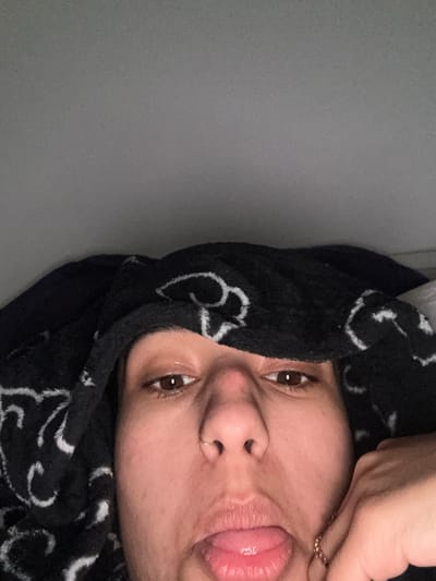 A close-up, angled shot features a person's face. The individual has brown eyes, a nose piercing, and is sticking their tongue out slightly. A dark, patterned fabric, possibly a towel or headscarf, frames their face. One hand with a ring on the finger touches the side of the face. The background is a plain gray wall.