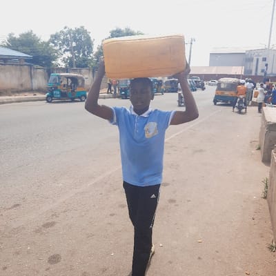 The image shows a young boy wearing a blue polo shirt and black pants walking down the street in Kufang, Nigeria. He is carrying a large yellow plastic container on his head. The container appears to be filled with water. There are several three-wheeled vehicles parked on the side of the road. The boy is walking away from the camera toward a concrete barrier on the right. There is a building with a tin roof in the background. The road is paved and appears to be in need of repair. The location is a residential area with trees and shrubs on the sides of the road. The coordinates (latitude: 9.863, longitude: 8.867) place this location in Kufang, Nigeria.