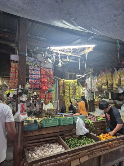 The image depicts a small general store, likely located in Parañaque, Philippines. Several individuals are visible, including one man in a black t-shirt examining vegetables (cucumbers, tomatoes, onions) displayed in wooden crates along the right edge of the frame. A woman in an orange shirt is seen further into the store, possibly serving or stocking items. Shelves and hanging displays contain a variety of goods, including instant noodles, snacks, and household items. The store's structure appears to be basic, with corrugated metal roofing and exposed wiring. Goods are organized and displayed in a way to maximize space in a densely packed arrangement. Green plastic crates can be seen among the shelves and display. The store sells fresh produce like garlic, calamansi, onion.